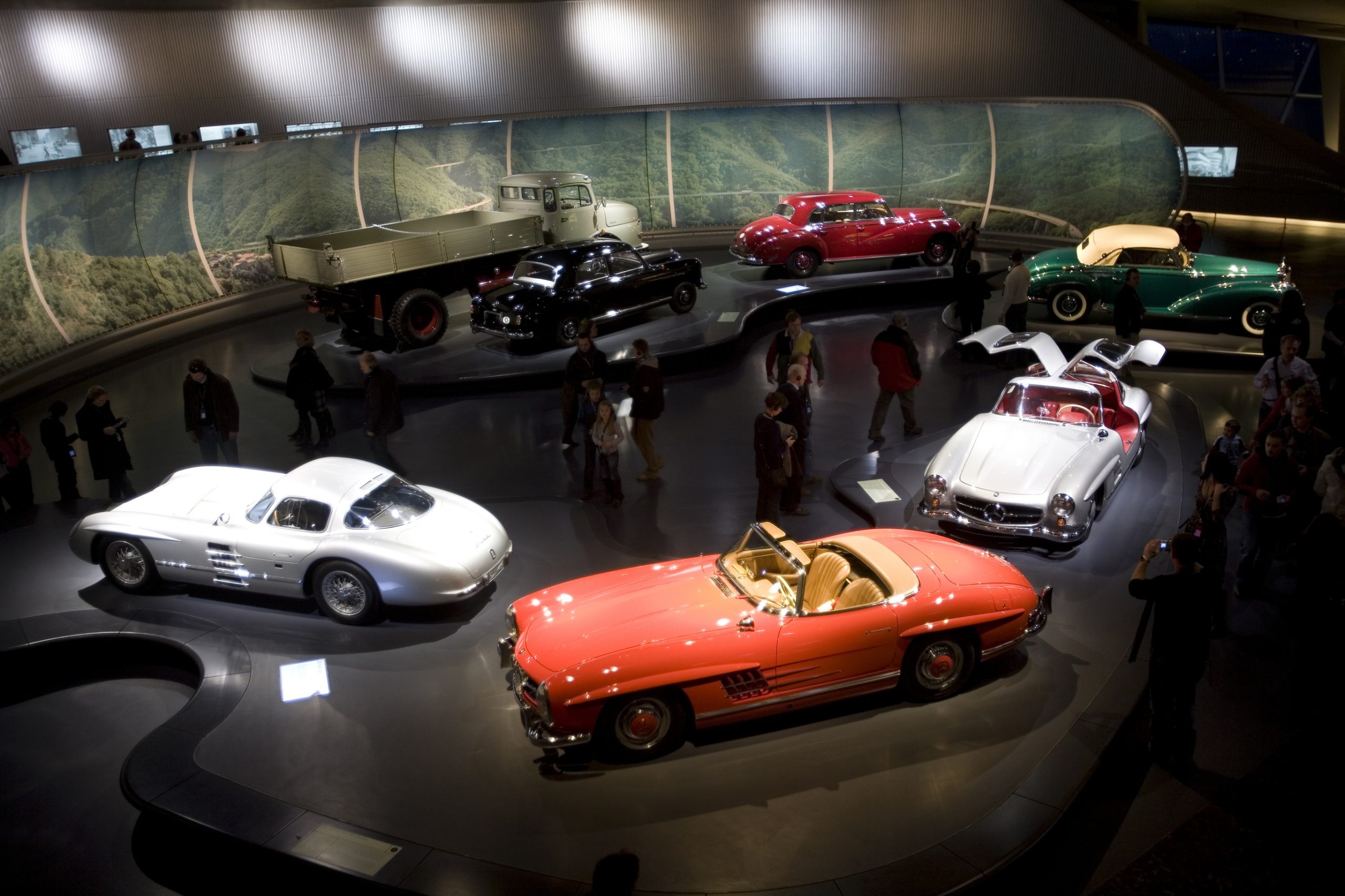 Model cars under a light at a museum with pedestrians looking at them.