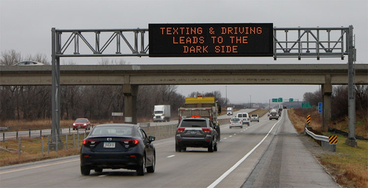 Cars on an interstate driving underneath a digital sign that says "Texting & Driving Leads to the Dark Side."