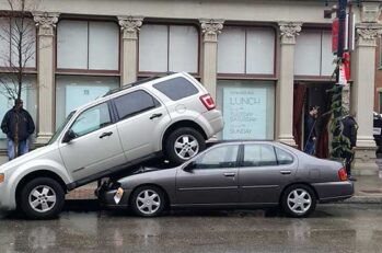 White SUV parked on top of a small grey 4-door car.