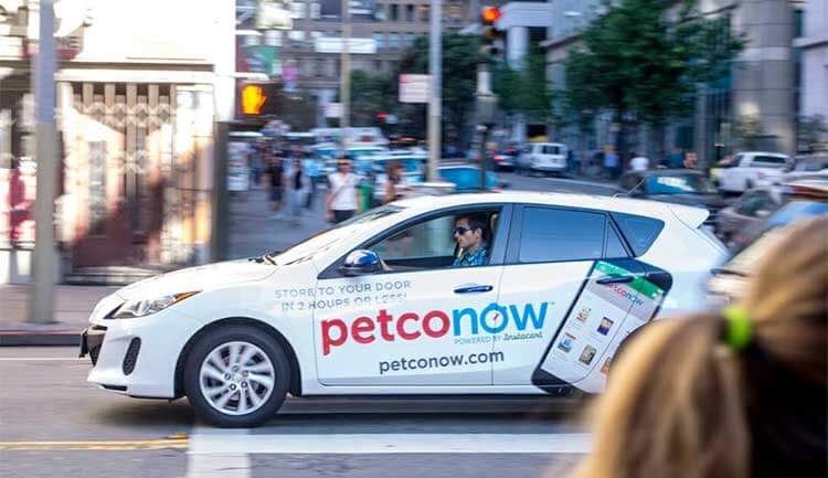 White car driving through a cross walk that reads "petco now" on the side of it.