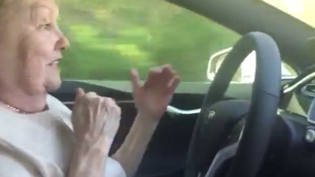 An elderly lady driving a car with her hands off of the steering wheel.