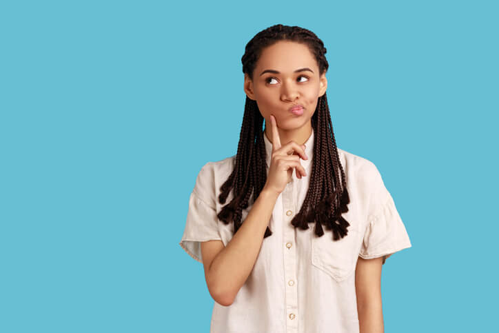 Portrait of a thoughtful woman with black dreadlocks who keeps her index finger on cheek, considers something, has a pensive expression, wearing white shirt. Indoor studio shot isolated on blue background.