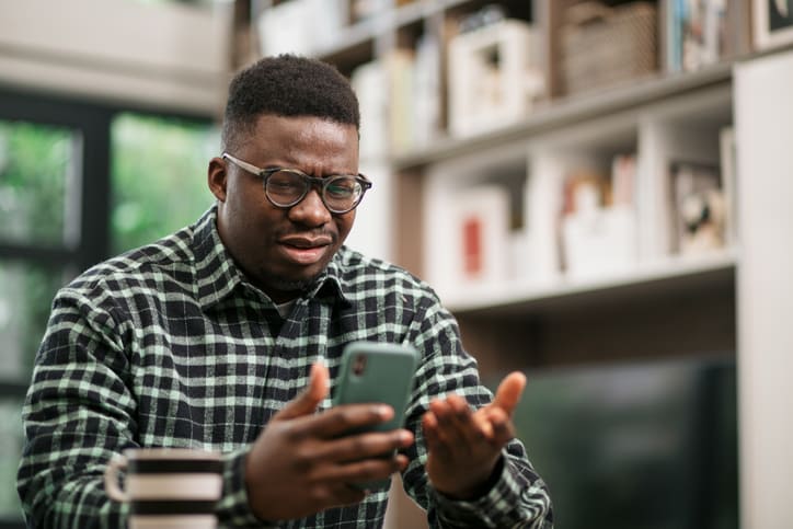 Annoyed young African American man sitting at home, reading some bad news on his smart phone using a mobile app and expressing his displeasure