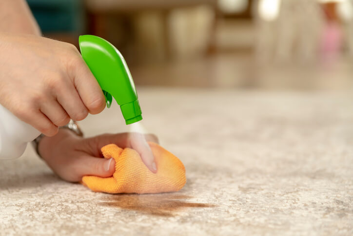 A bottle of cleaner is sprayed above a hand with a sponge clearing a surface.