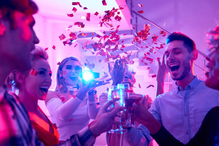 Group of happy festive people enjoying holiday celebration at private house party raising glasses under bursts of confetti