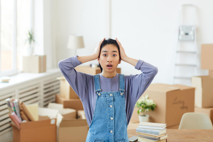 Waist up portrait of young Asian woman panicking while standing among cardboard boxes in empty room and looking at camera with big eyes after moving.