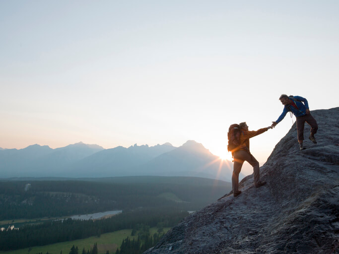 The sun is rising over the distant peaks as one mountain climber extends a hand to help another below them.